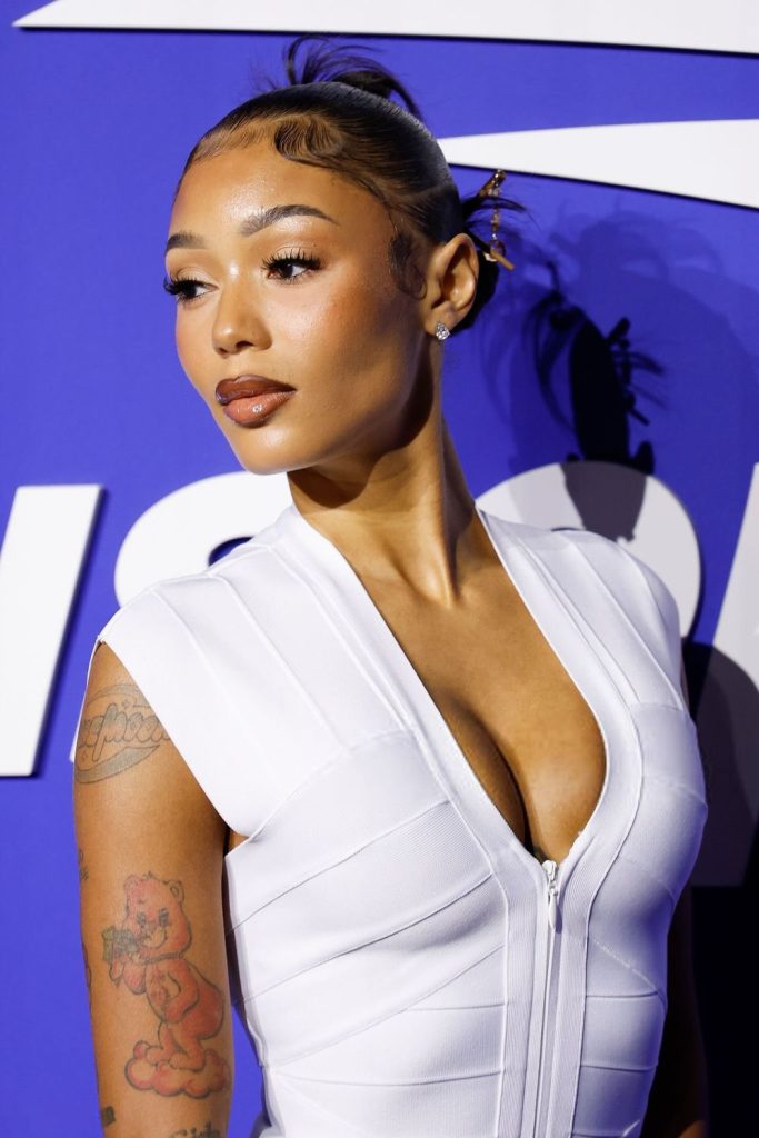 A woman wearing a white zip-up dress poses in front of a blue and white background. She has her hair styled up with decorative pins and has visible tattoos on her arm, including a red bear.
