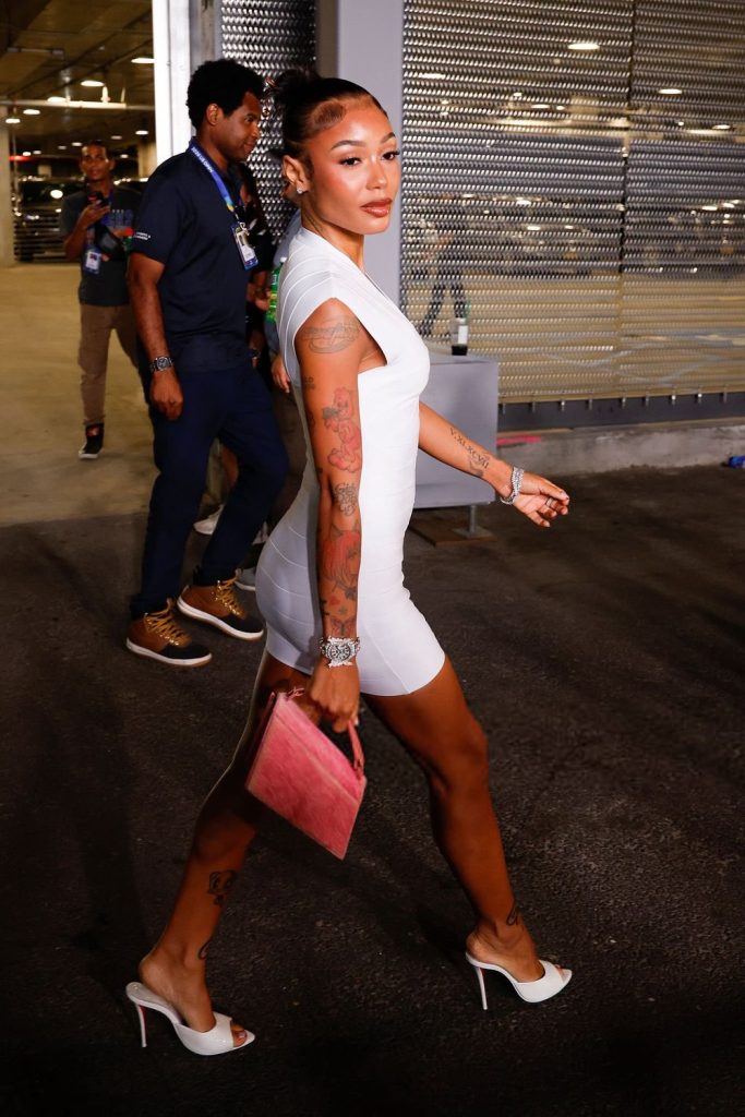Woman in a fitted white dress and heels walks outside at night, holding a pink handbag. She has tattoos on her arms and legs. Other people and a lit building are visible in the background.