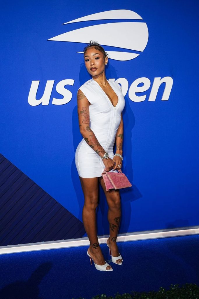 A woman in a short white dress and white heels stands in front of a blue US Open backdrop, holding a pink clutch. She has tattoos on her arms and legs and wears jewelry.