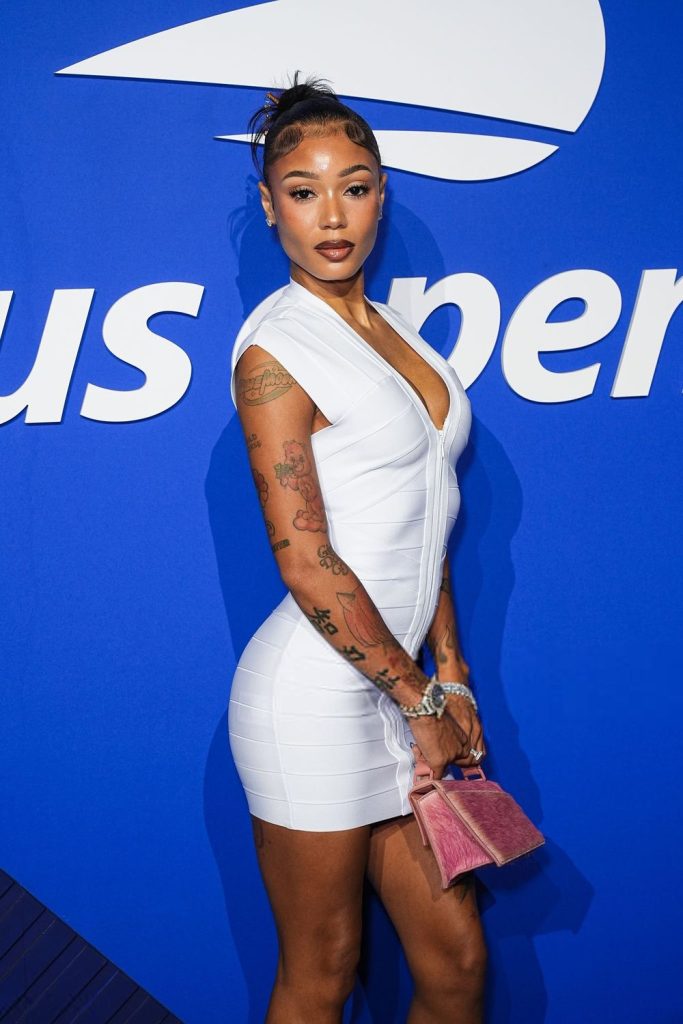 A woman in a fitted white dress poses on a blue carpet, holding a small pink purse. She has visible tattoos on her arms and styled hair, standing in front of a blue background with white text.