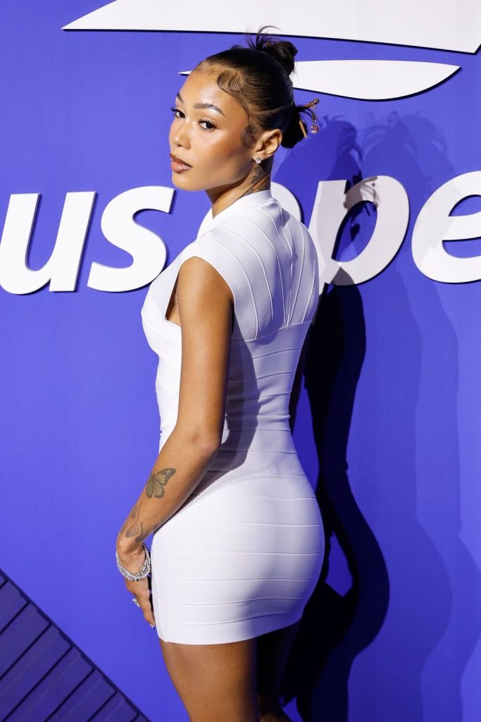 Woman in a white fitted dress poses sideways in front of a blue wall with white text. She has a tattoo on her arm, hair in a bun, and looks over her shoulder toward the camera.