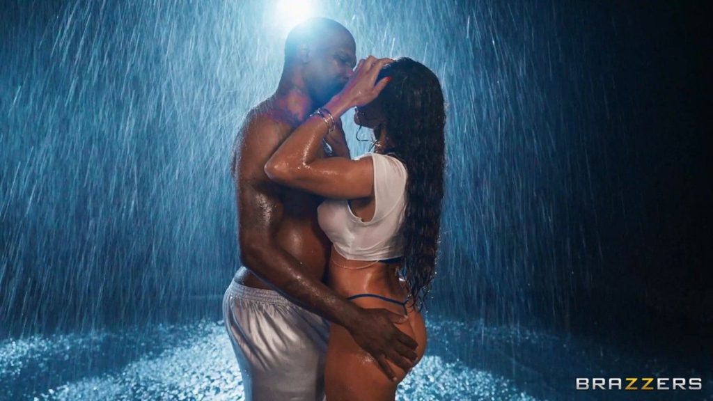 Woman with long dark curly hair in a wet white crop top and blue bikini bottoms embracing and kissing a bald muscular man under heavy rain.