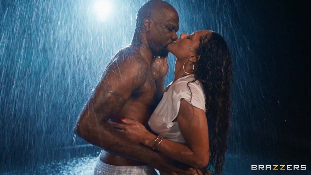 Woman with long dark curly hair in a wet white t-shirt kissing a bald muscular man under heavy rain, his hand on her face.