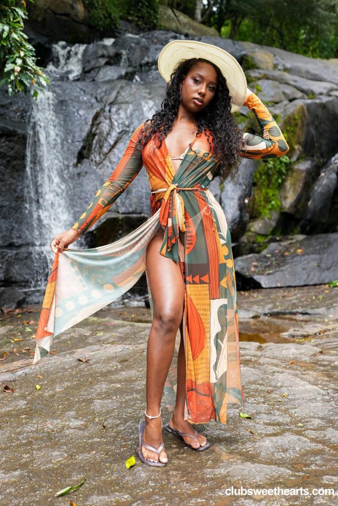 Woman with curly dark hair wearing a wide-brimmed straw hat, patterned long-sleeve cover-up, and bikini, standing in front of a waterfall while holding the cover-up open.