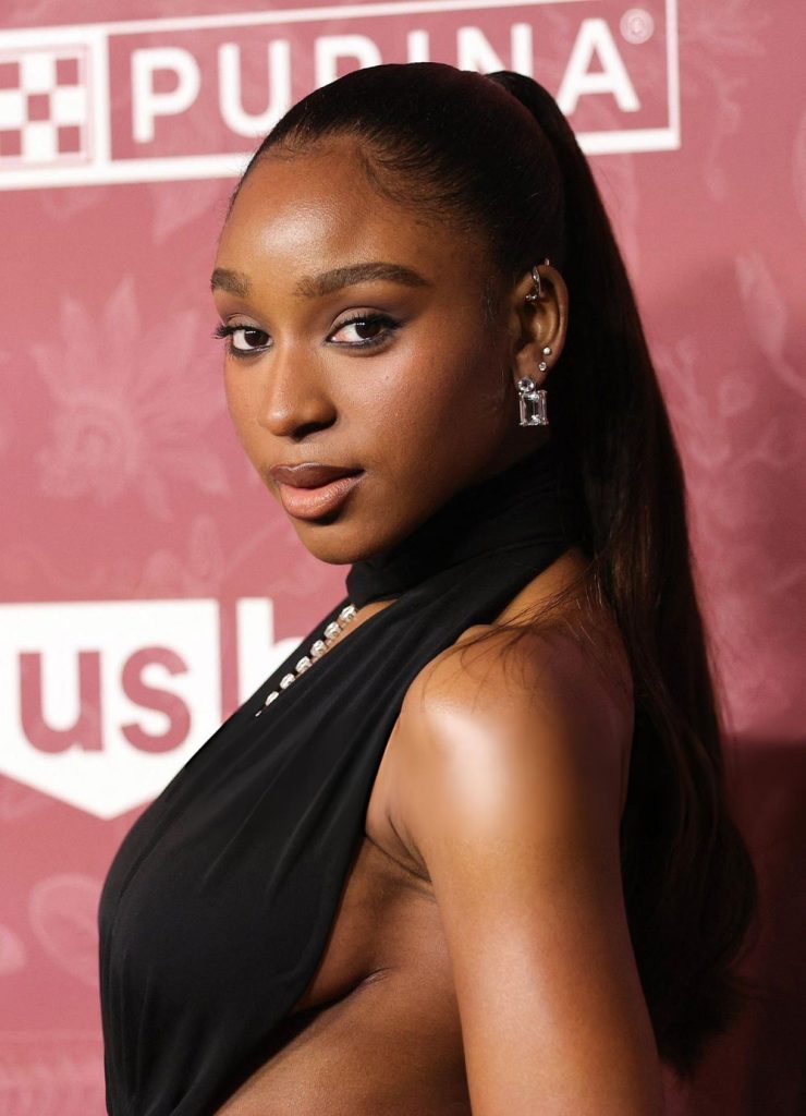 Close-up of a woman with dark hair in a high ponytail against a branded backdrop, wearing a black halter-style top with a jeweled detail, looking toward the camera.