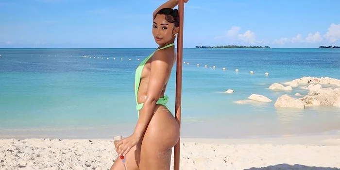 Black woman in a lime green swimsuit stands on a beach under a white umbrella, holding a glass.