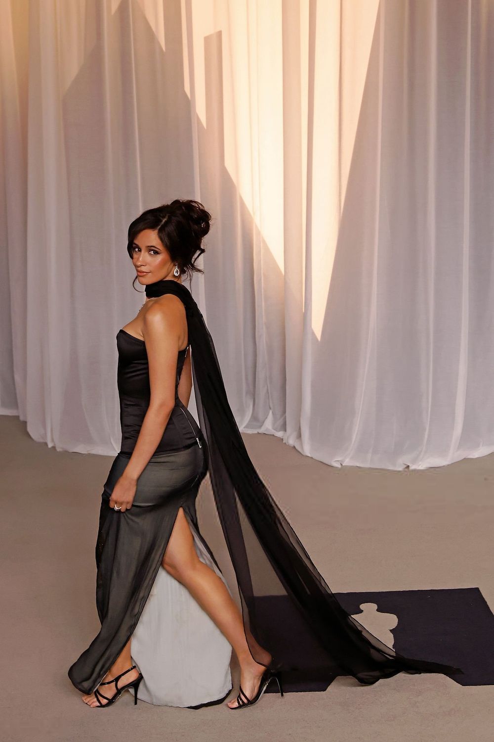 Woman with dark hair styled in an updo, wearing a black strapless corset top, sheer dark skirt with a high slit, black choker, and long sheer black fabric trailing behind, standing in three-quarter view looking back over her shoulder while wearing black strappy high-heeled sandals.