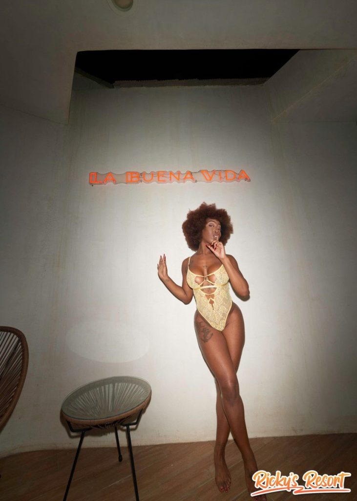 Woman with curly brown hair in a yellow lace cut-out bodysuit standing indoors against a wall with a red “LA BUENA VIDA” neon sign, one hand near her face, facing the camera.