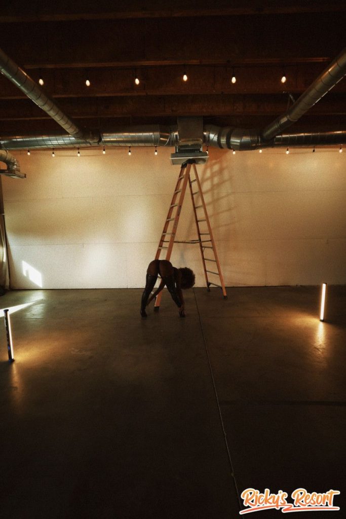 Woman with curly brown hair wearing black thigh-high boots and a dark outfit, bent over in a large empty indoor space with a tall ladder and string lights.