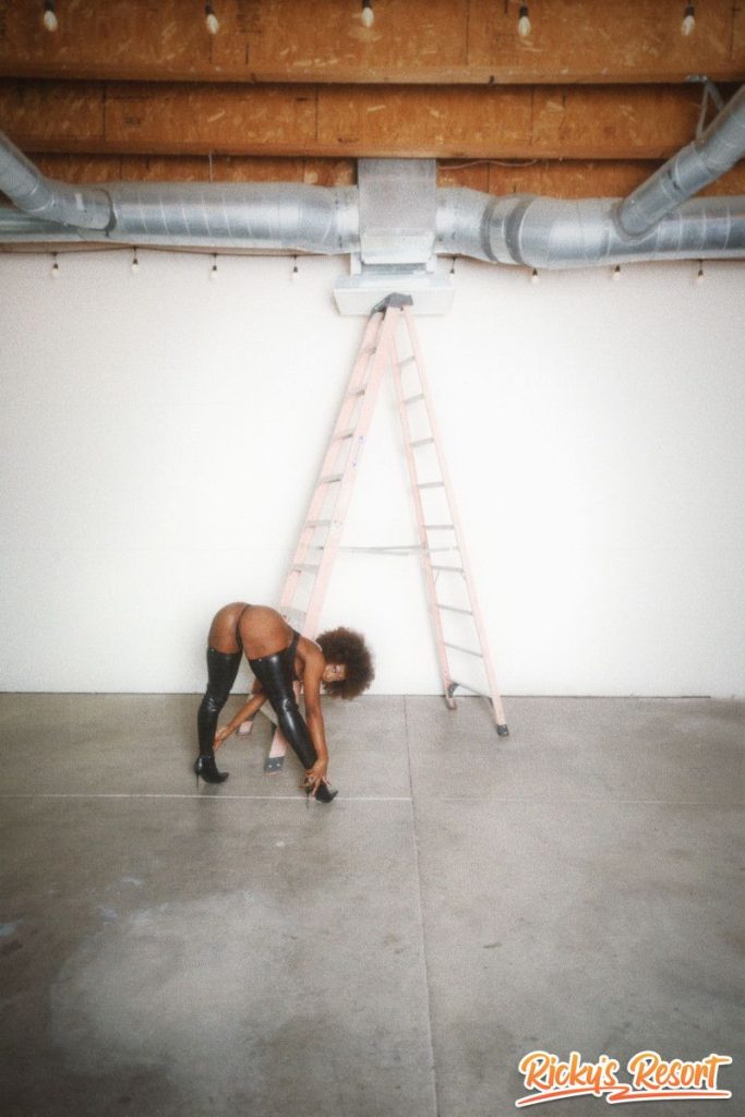 Woman with curly brown hair in black thigh-high boots and a dark outfit, bent over near a tall ladder in a large empty indoor space with string lights.