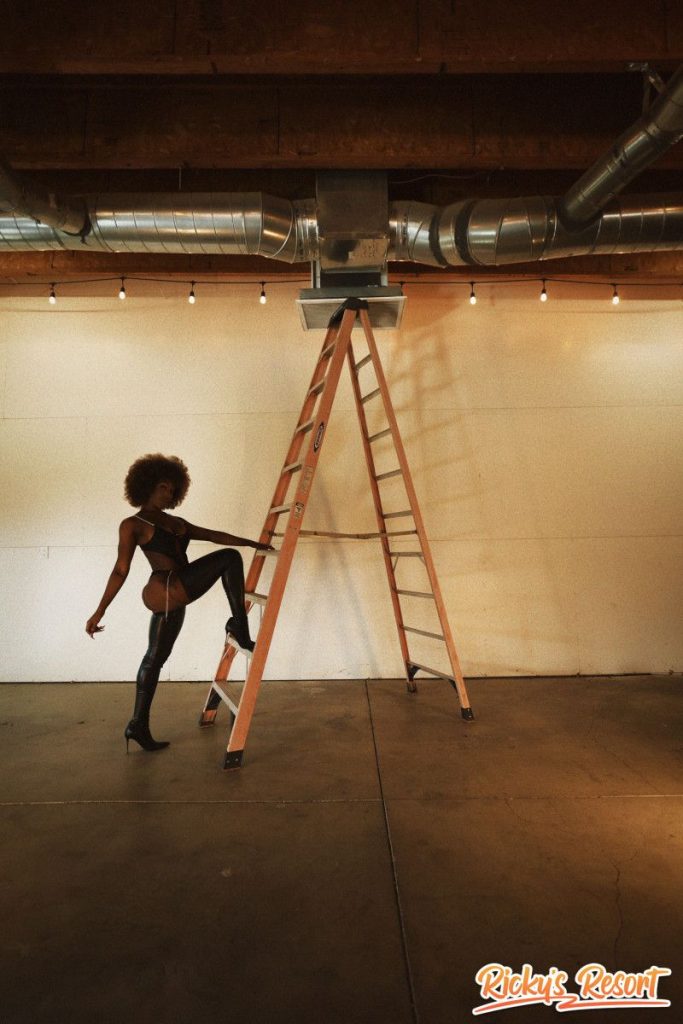 Woman with curly brown hair in a black cut-out bodysuit and thigh-high boots, standing with one leg raised on a tall ladder in a large empty indoor space.