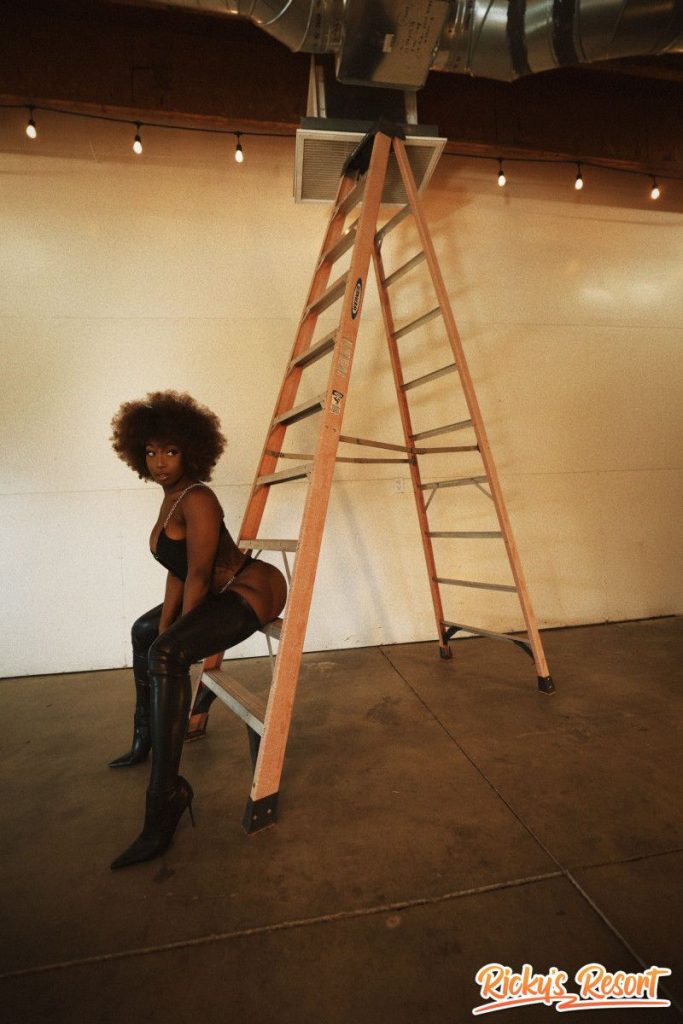 Woman with curly brown hair in a black cut-out bodysuit and thigh-high boots, seated on the lower rungs of a tall ladder in a large empty indoor space, looking at the camera.