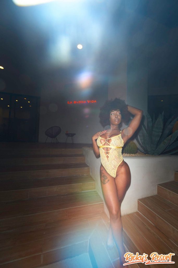 Woman with curly brown hair in a yellow lace bodysuit standing on outdoor steps, one hand near her head, facing the camera under bright light at night.