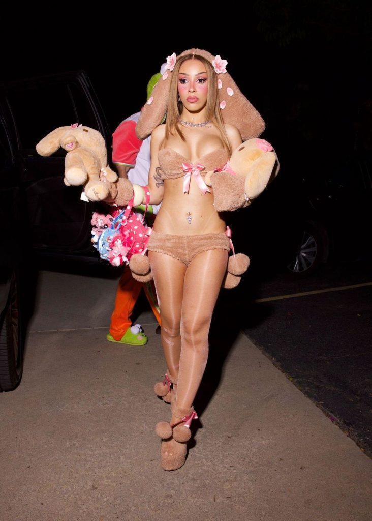 Doja Cat wearing a brown bunny-themed outfit with plush bunny ears, carrying two stuffed rabbits and a patterned bag.