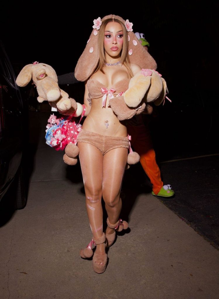 A person dressed in a fuzzy brown rabbit costume with pink bows, holding two stuffed rabbits and a small bouquet.