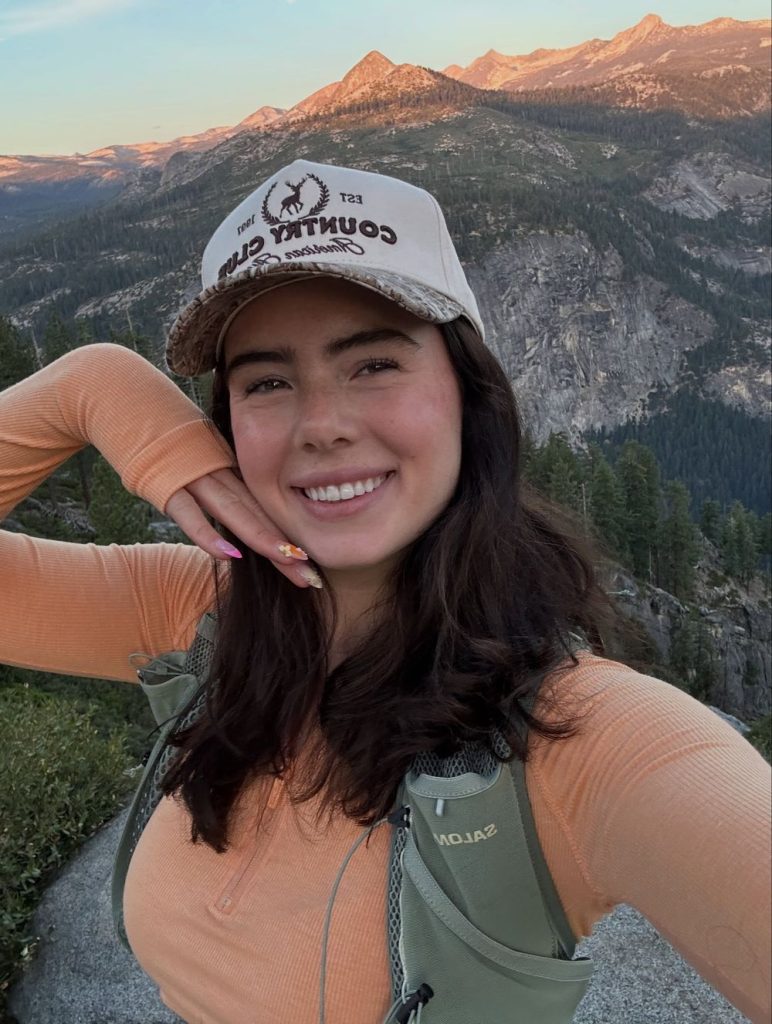 Woman with long dark hair wearing a beige baseball cap and peach long-sleeve top, smiling in a close-up selfie with a mountain landscape in the background.