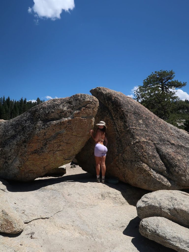 Woman wearing a wide-brimmed hat and light purple shorts, standing with her back to the camera between two large boulders under a clear blue sky.
