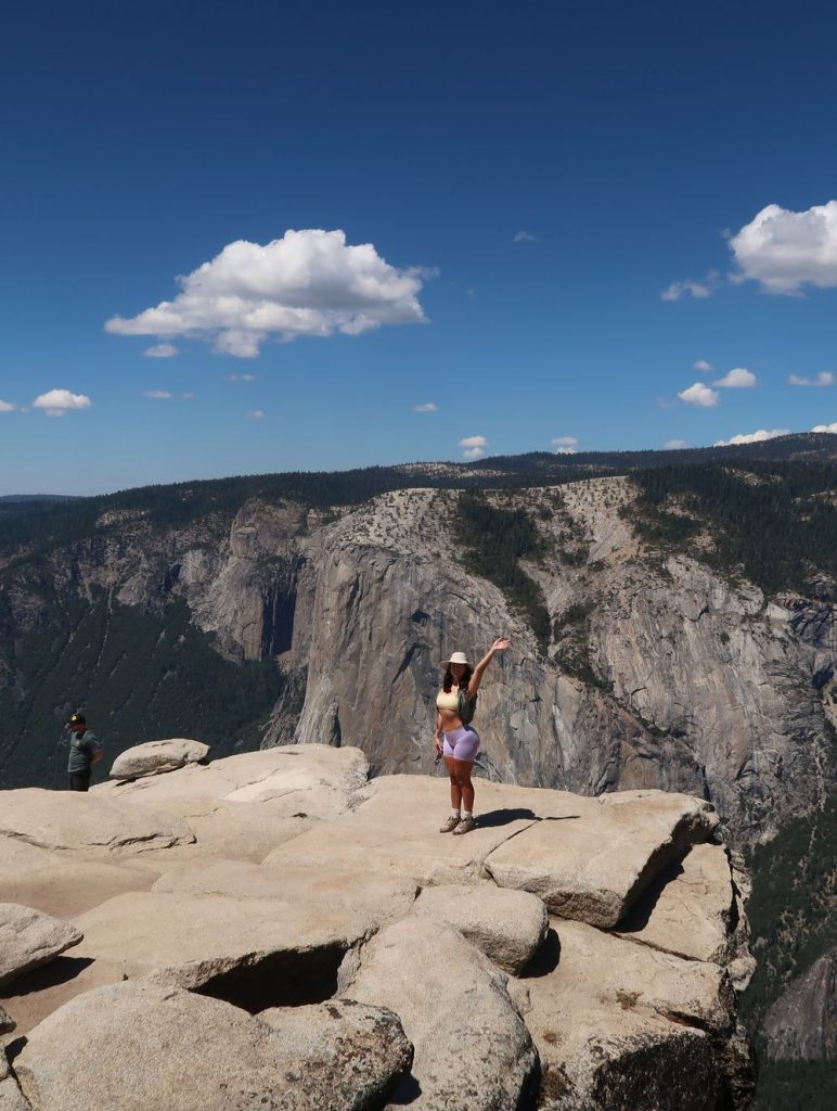 Woman wearing a wide-brimmed hat, yellow top, and light purple shorts, standing on a rocky overlook with one arm raised and a large granite formation in the background.