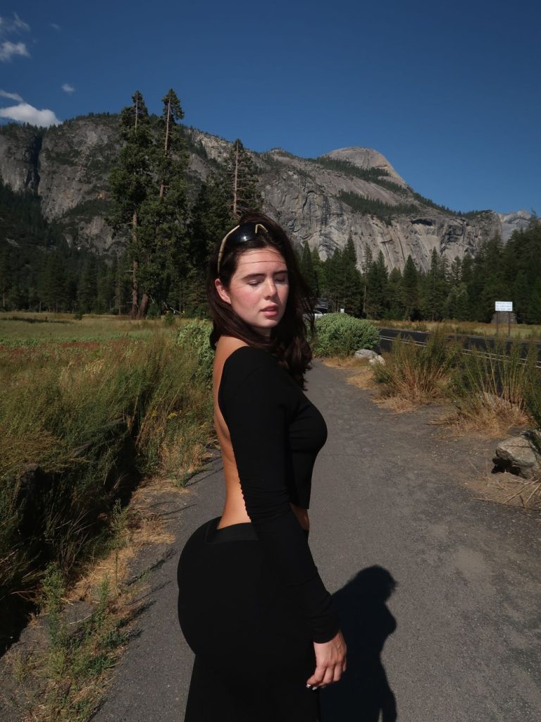 Woman with long dark hair wearing a black long-sleeve open-back top and black pants, standing on a paved path with a mountain landscape and trees in the background.