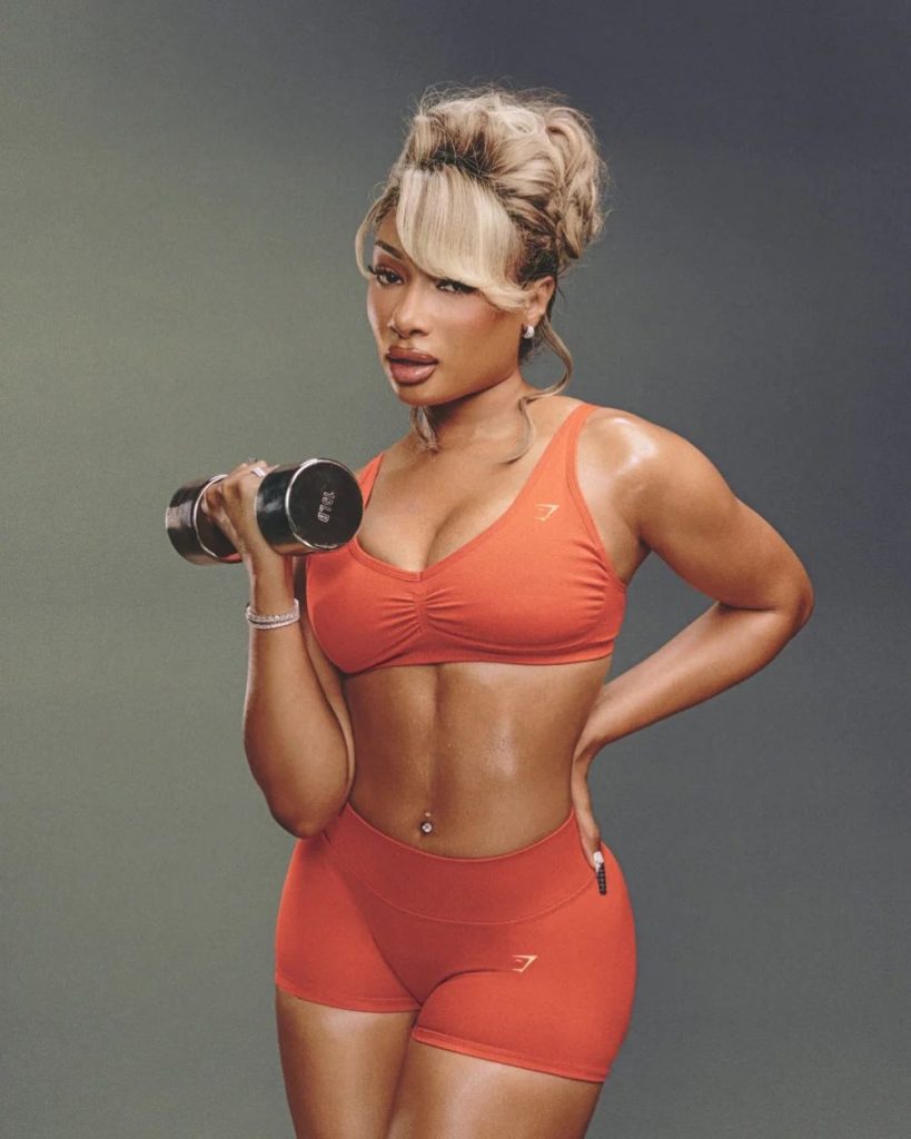 Megan Thee Stallion wearing a matching orange sports bra and shorts lifts a dumbbell.