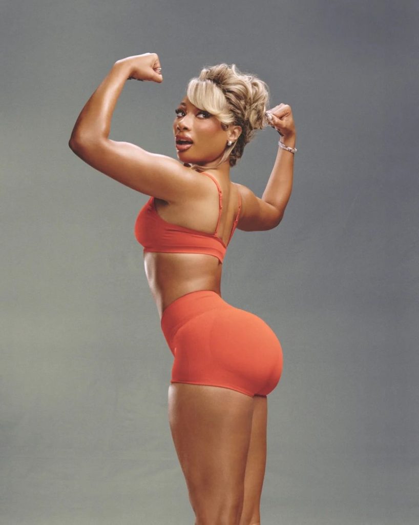 Megan Thee Stallion wearing a matching orange sports bra and shorts while flexing her arm muscles.