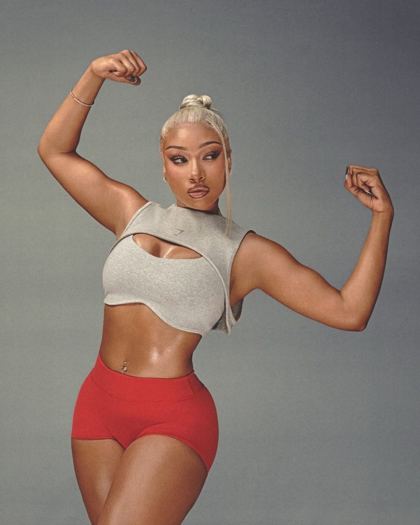 Megan Thee Stallion posing with her arms flexed, wearing a grey cropped tank top and red shorts.
