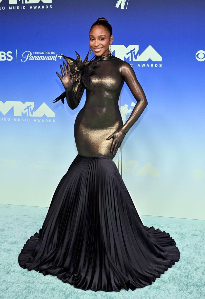 Normani poses on the MTV Video Music Awards red carpet wearing a metallic gold long-sleeved bodysuit with a black floral shoulder accent and a floor-length pleated black skirt.