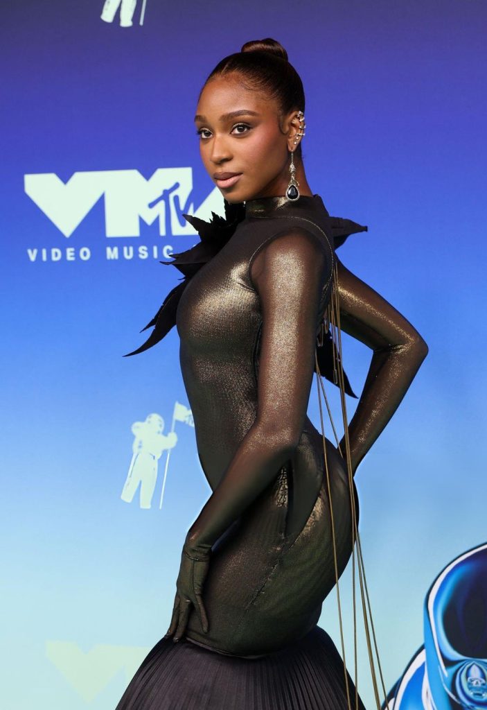 Normani posing on a blue MTV VMA red carpet backdrop wearing a metallic, long-sleeved, high-neck gown with dark feather accents.