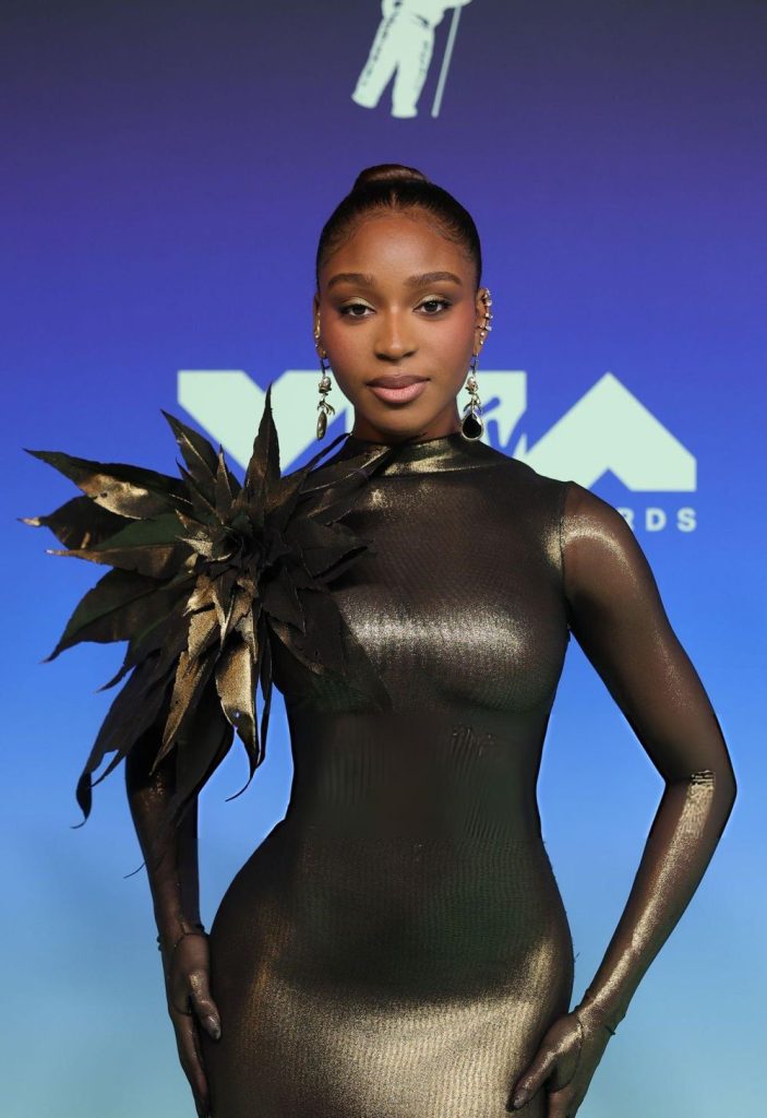 Normani poses on a blue MTV VMA red carpet wearing a metallic gold long-sleeved bodysuit with a large, dark flower detail on the shoulder.