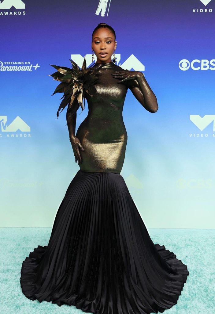 Normani posing on a blue carpet in a high-neck, metallic gold long-sleeved gown with a large black floral shoulder accent and a pleated black mermaid skirt.
