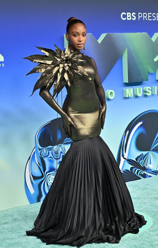 Normani poses on a blue carpet in a form-fitting metallic gold gown with a large sculptural floral shoulder piece and a floor-length black pleated skirt.
