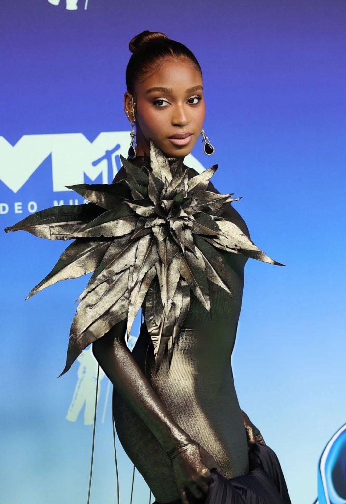 Normani posing on a blue carpet in a metallic gown featuring a large, star-shaped embellishment on the shoulder.