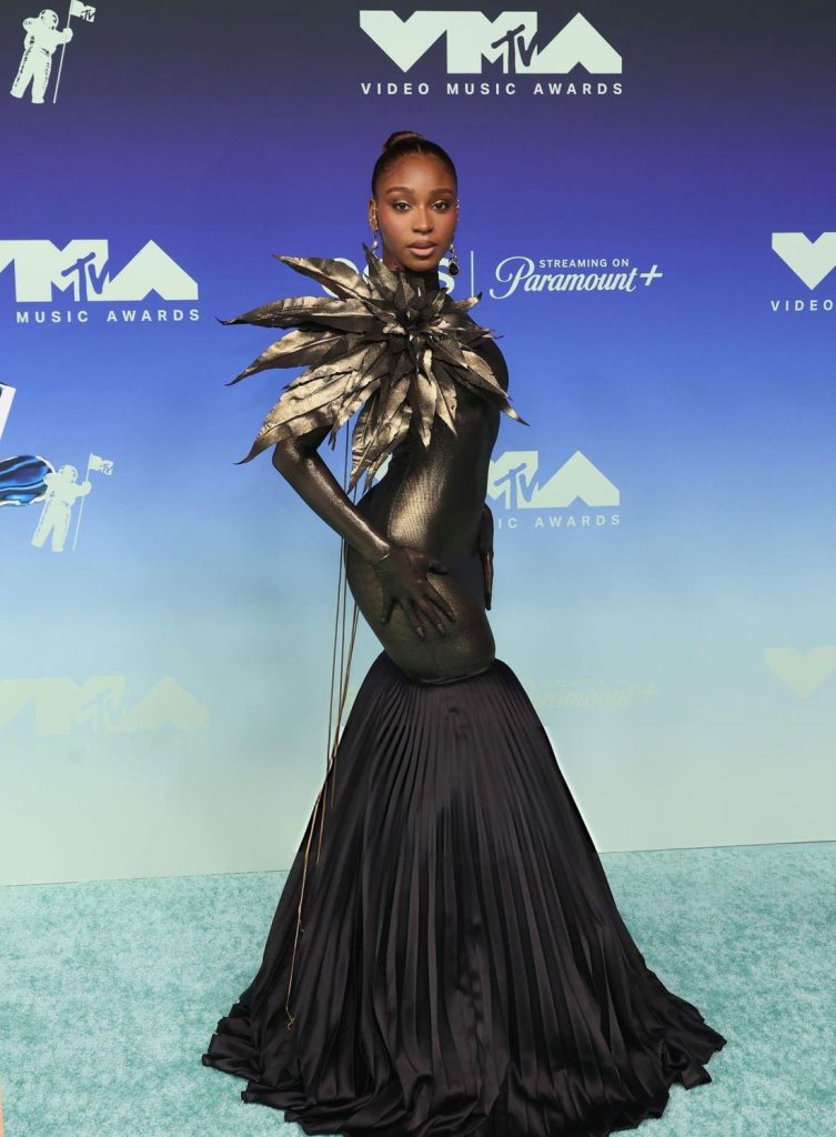Normani posing on the MTV Video Music Awards red carpet wearing a black floor-length gown with a sculptural metallic shoulder accent.