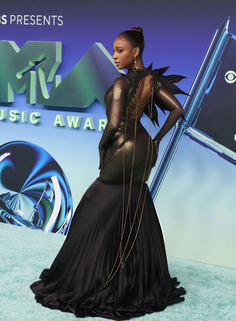 Normani poses on the MTV Video Music Awards red carpet wearing a backless gold and black gown with a long pleated skirt and thin golden chains draping down her back.