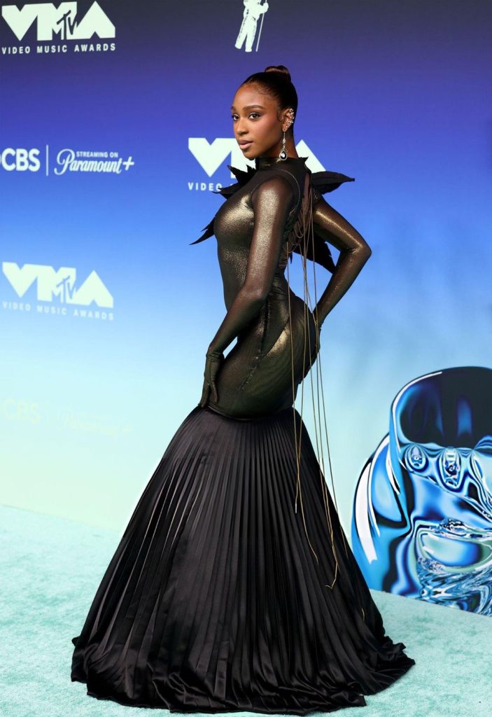 Normani poses on a blue carpet wearing a high-necked, metallic gold bodysuit with long sleeves, gloves, and a pleated black floor-length skirt at the MTV Video Music Awards.
