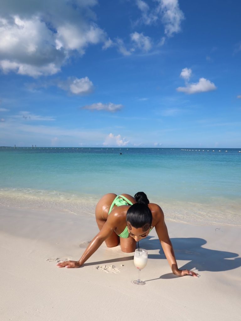 A woman in a neon green bikini kneeling on a sandy beach to sip a cocktail from a glass.
