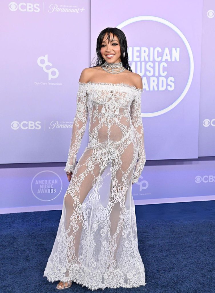 Tinashe poses on a blue carpet in front of an American Music Awards backdrop wearing an off-the-shoulder white lace gown and a layered crystal choker.