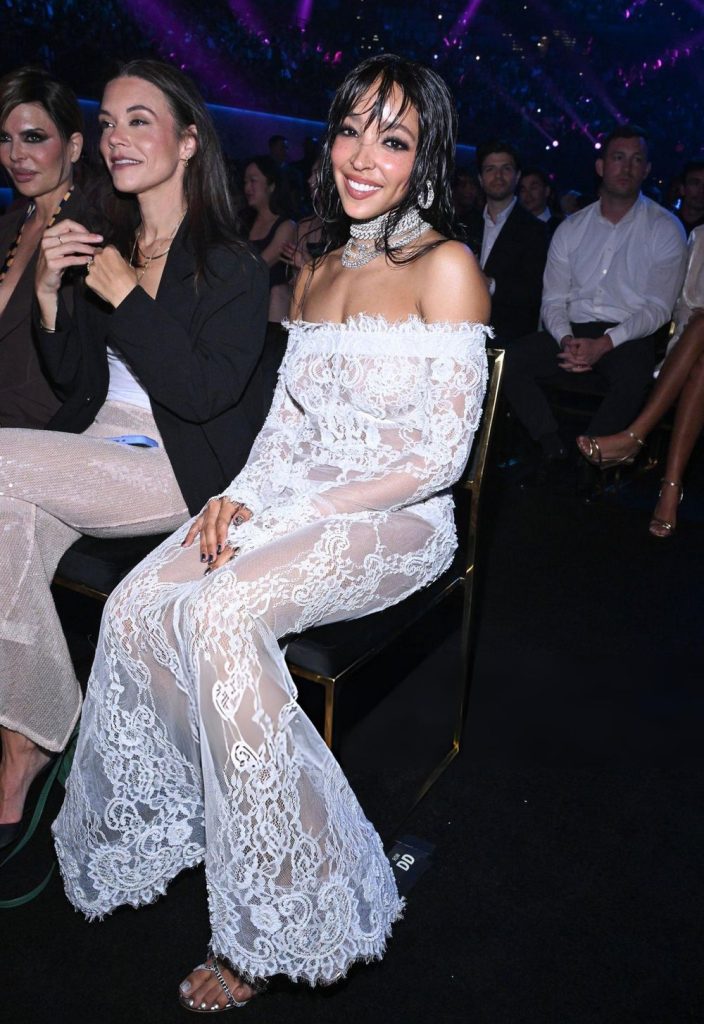 Tinashe smiling while wearing a long-sleeved, off-the-shoulder white lace gown at an event.